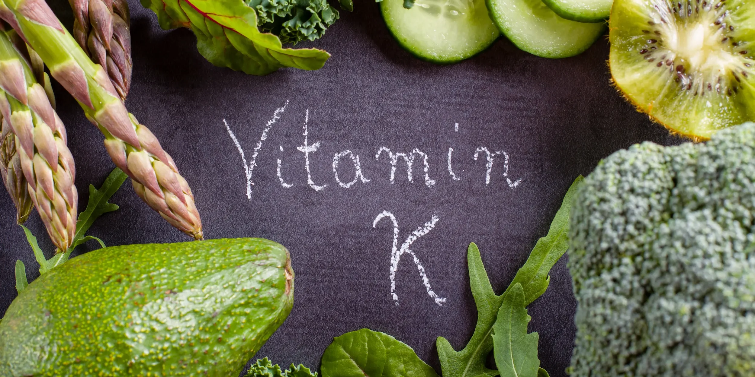 Vitamin K’ written in chalk on slate, surrounded by various food sources of the vitamin.