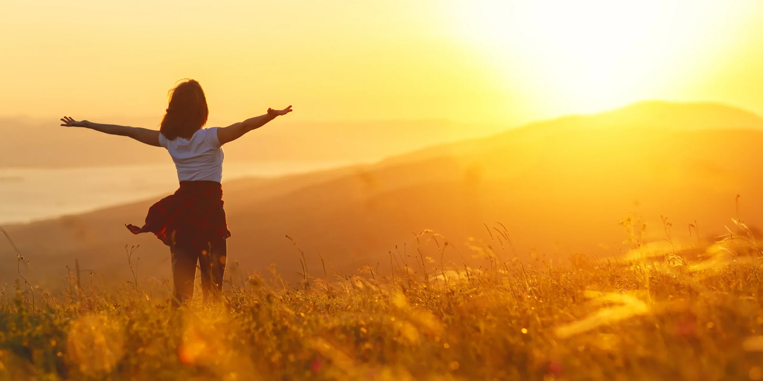 A woman stands in a sunny field, spreading her arms wide and facing the sunset.