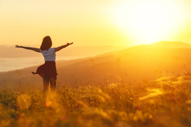 A woman stands in a sunny field, spreading her arms wide and facing the sunset.