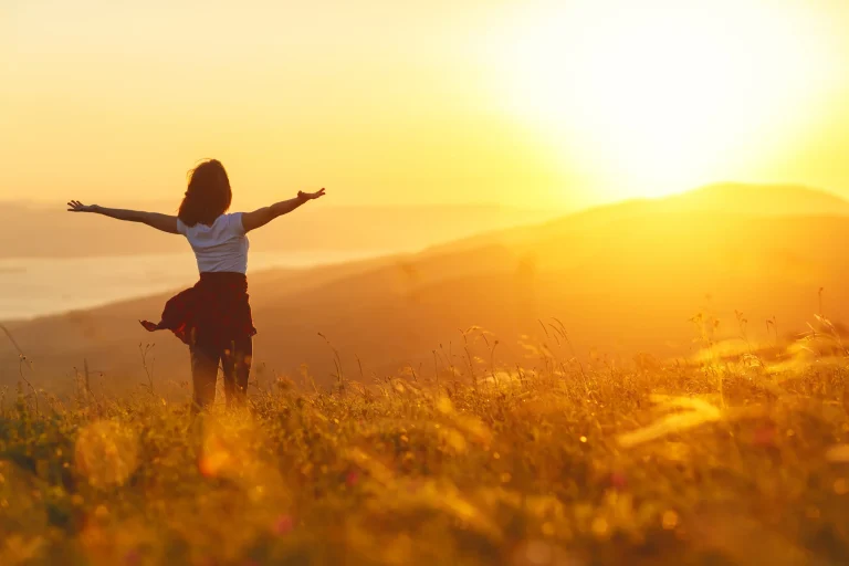 How do you get vitamin D A woman stands in a sunny field, spreading her arms wide and facing the sunset.