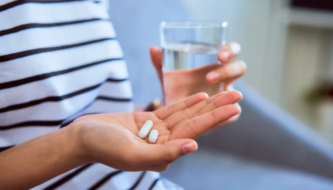 A woman holds two tablets in one hand and a glass of water in the other.