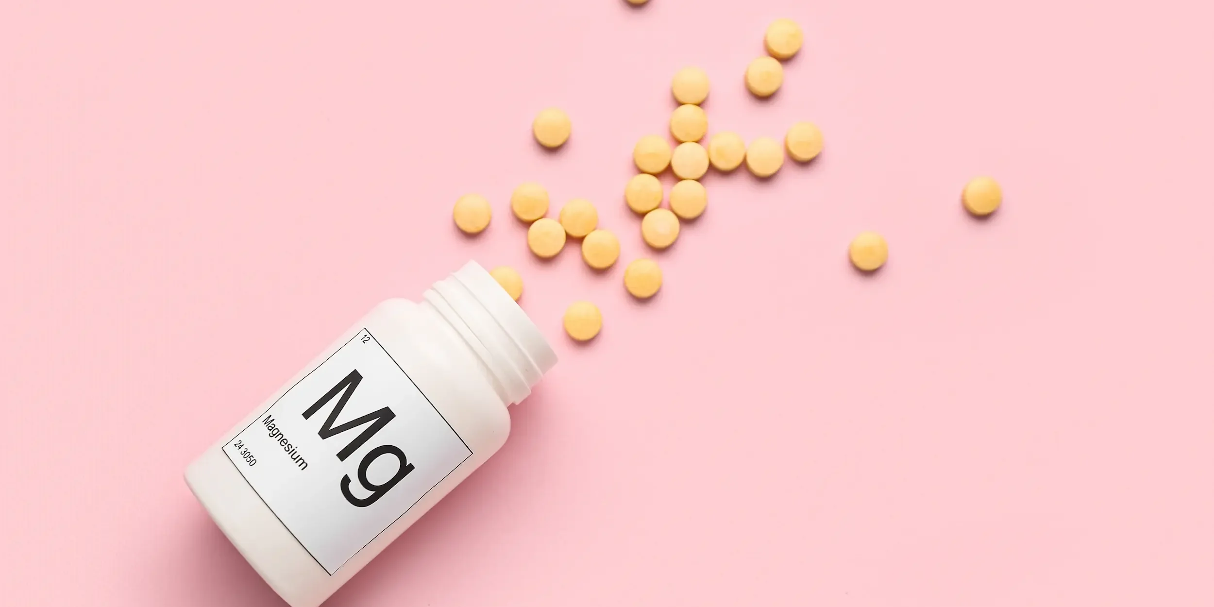 A bottle of pills spilling across a pink background. The bottle is labelled ‘Mg’ for magnesium.
