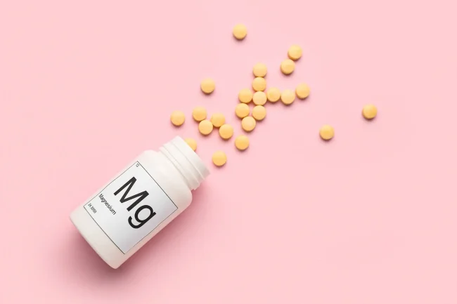 A bottle of pills spilling across a pink background. The bottle is labelled ‘Mg’ for magnesium.