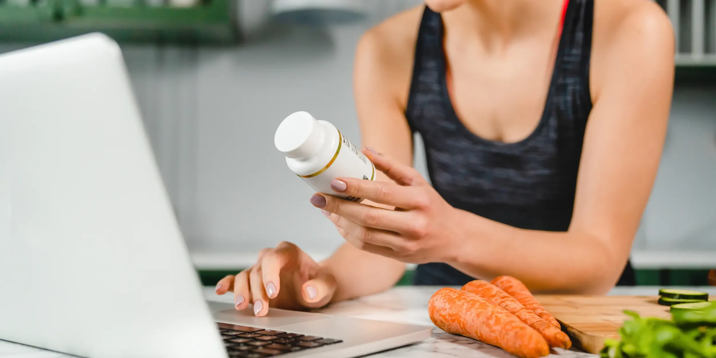 A woman in sports gear researches the ingredients of her food supplement on her laptop in the kitchen.