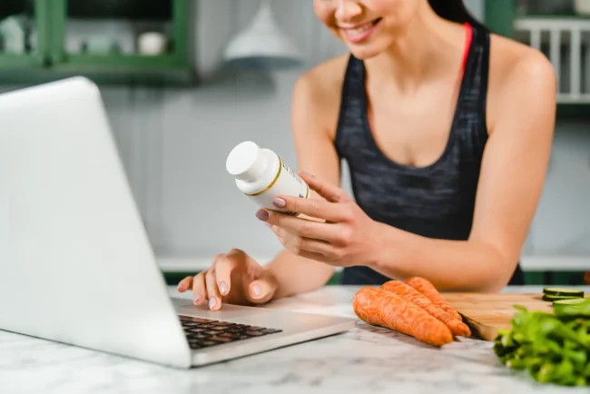 A woman in sports gear researches the ingredients of her food supplement on her laptop in the kitchen.
