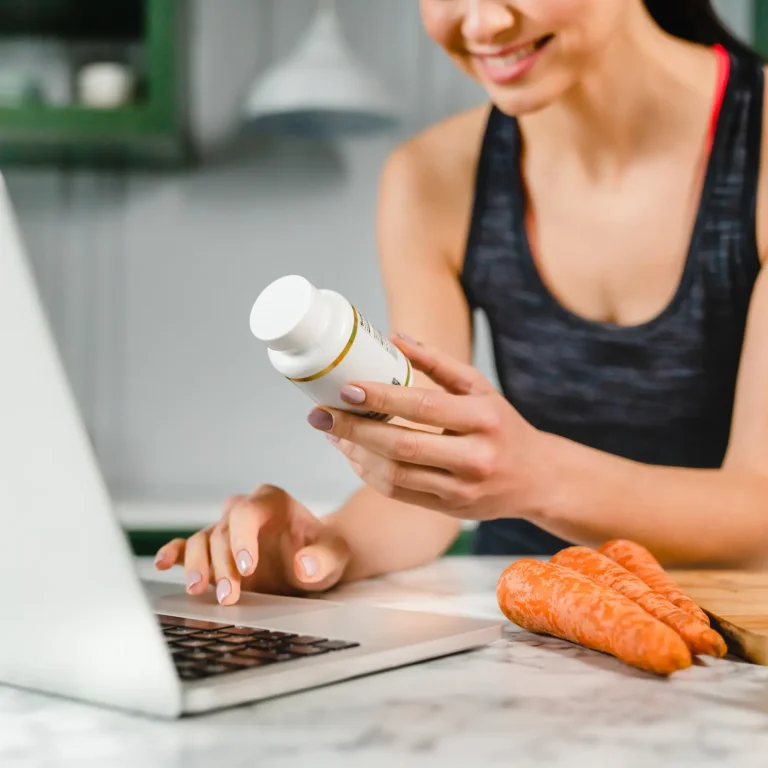A woman in sports gear researches the ingredients of her food supplement on her laptop in the kitchen.