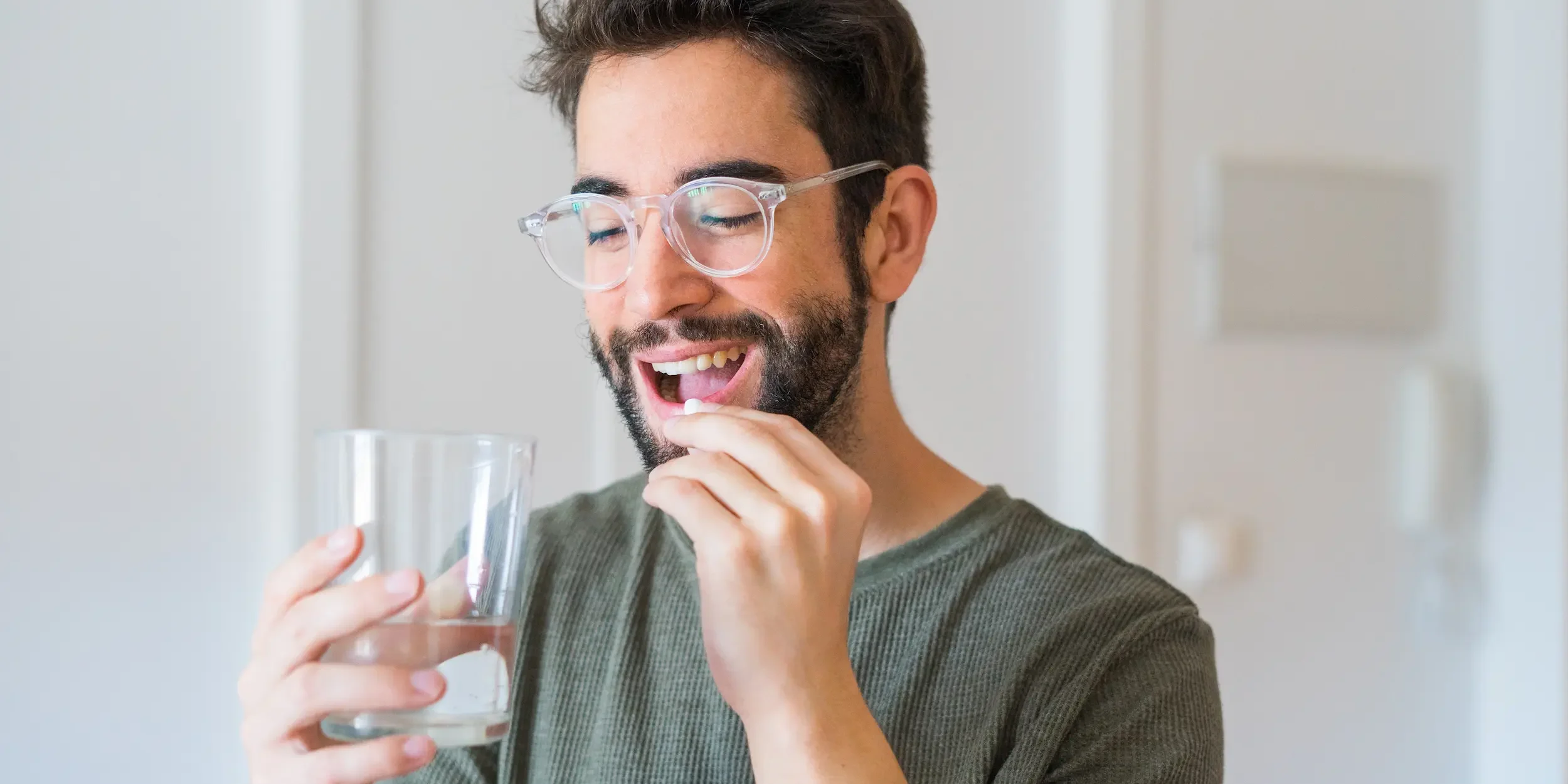 A man wearing clear-rimmed glasses takes a tablet with a glass of water.