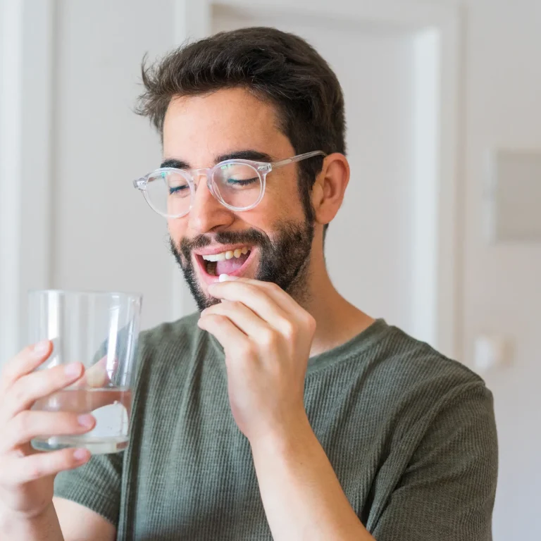 A man wearing clear-rimmed glasses takes a tablet with a glass of water.
