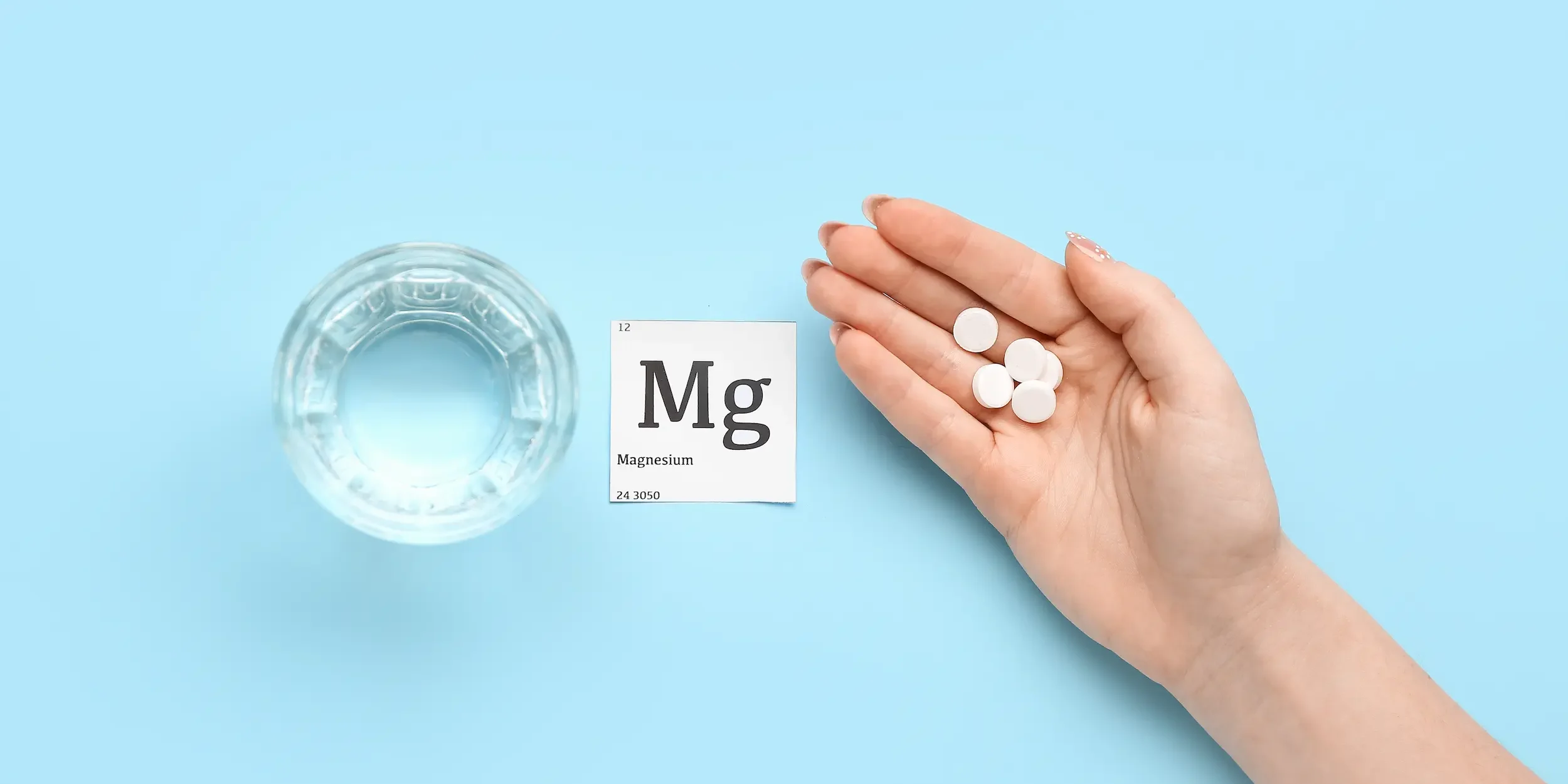 A glass of water and a square piece of paper reading ‘Mg Magnesium’ beside a hand holding several small pills, all on a light blue background.