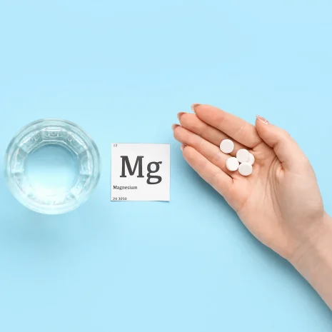 A glass of water and a square piece of paper reading ‘Mg Magnesium’ beside a hand holding several small pills, all on a light blue background.
