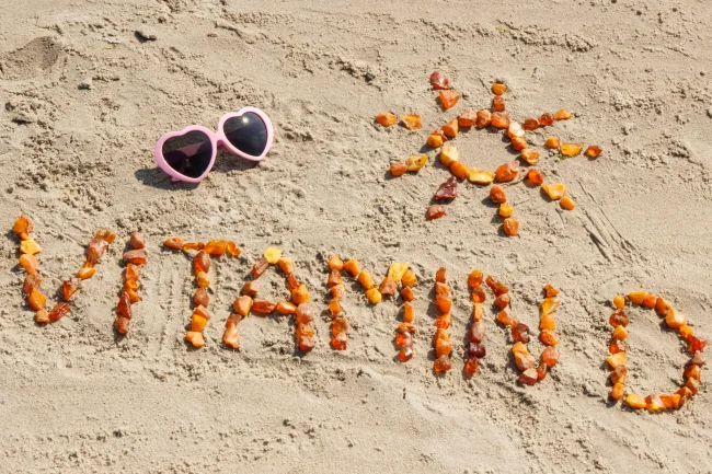 Amber sea glass spells out ‘Vitamin D’ and an image of a sun on a beach next to a pair of pink heart-shaped sunglasses.