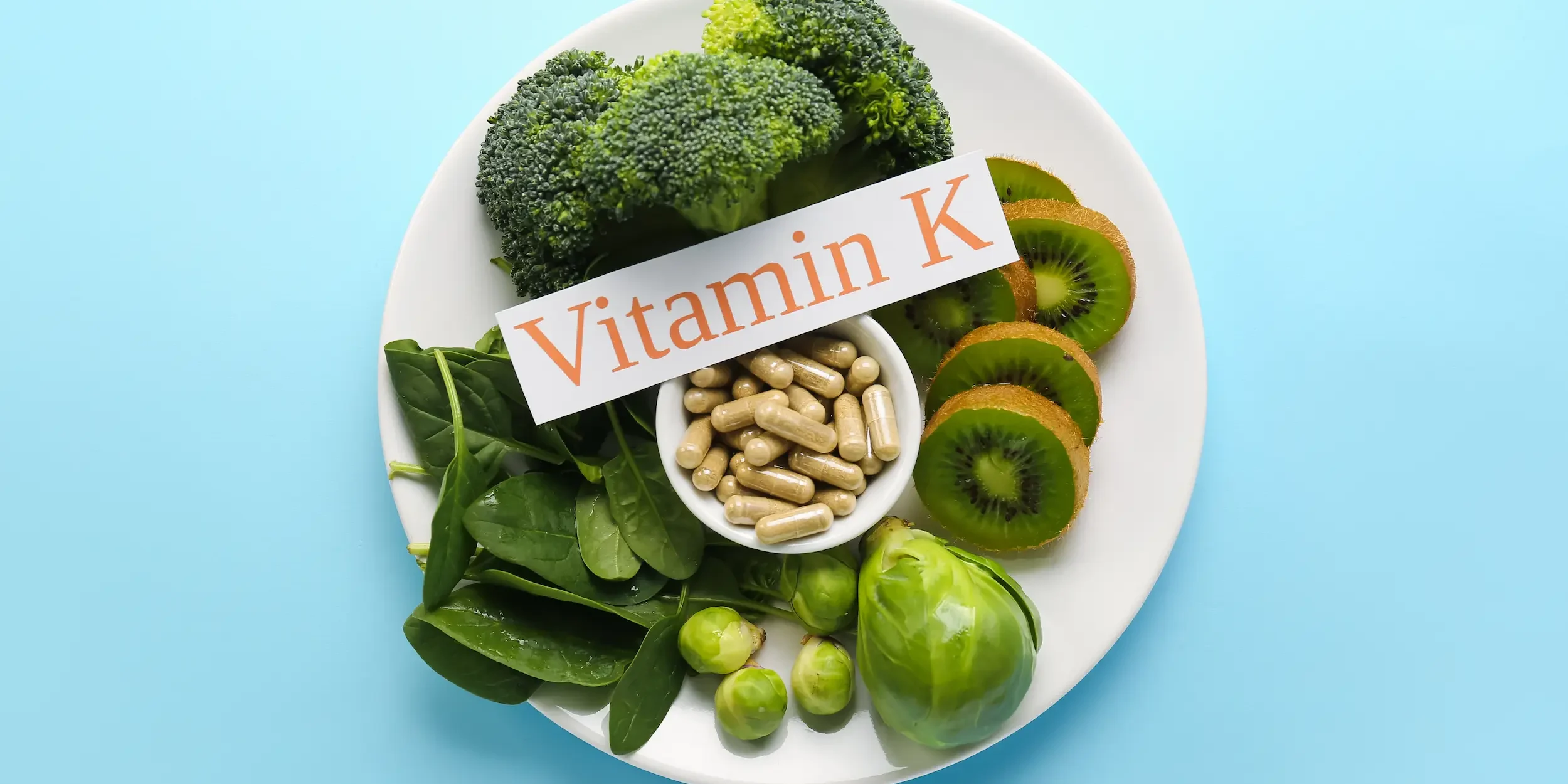 Are vitamin K supplements safe A plate of vitamin K-rich greens around a pot of tablets and a sign reading ‘Vitamin K’.