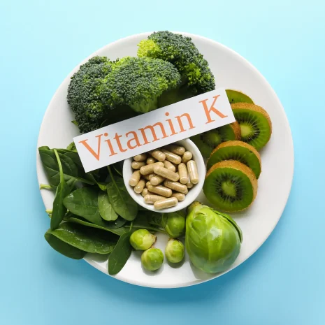 A plate of vitamin K-rich greens around a pot of tablets and a sign reading ‘Vitamin K’.