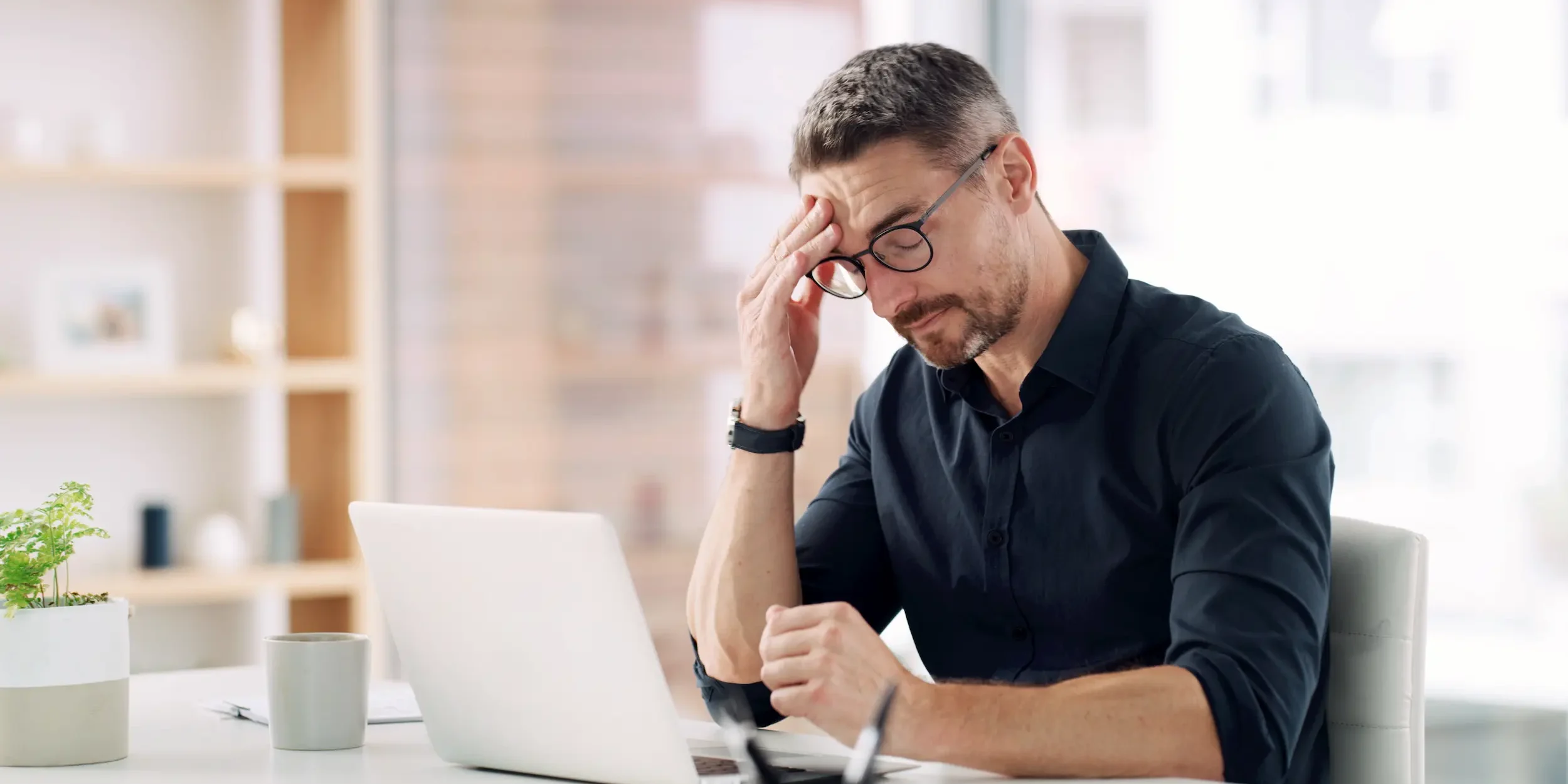 A man experiences a headache while working at a laptop in his office.