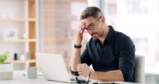A man experiences a headache while working at a laptop in his office.