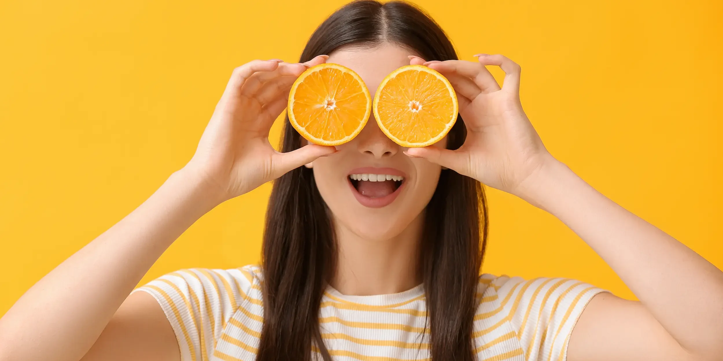 A woman holds two orange halves in front of her eyes, representing almost all of her required daily vitamin C intake.