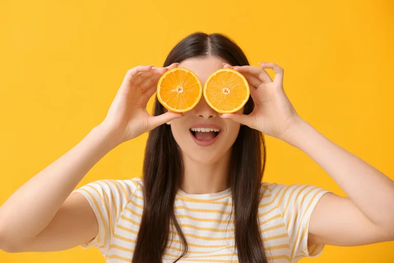 A woman holds two orange halves in front of her eyes, representing almost all of her required daily vitamin C intake.