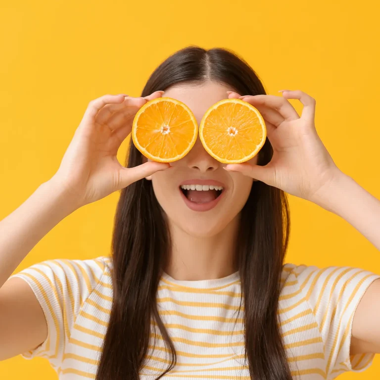 A woman holds two orange halves in front of her eyes, representing almost all of her required daily vitamin C intake.