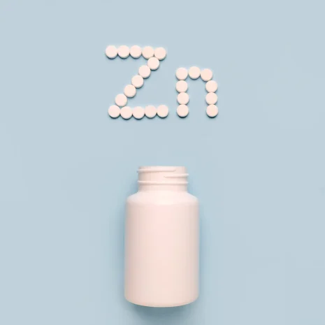 Small white pills spell out ‘Zn’ for zinc above a white pill bottle on a pale blue background.
