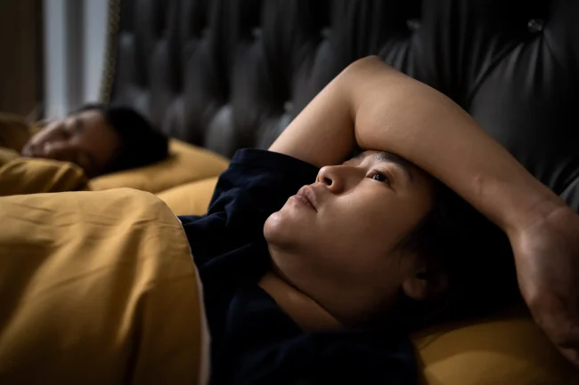 A woman lies awake in bed beside her sleeping husband, symbolising insomnia.