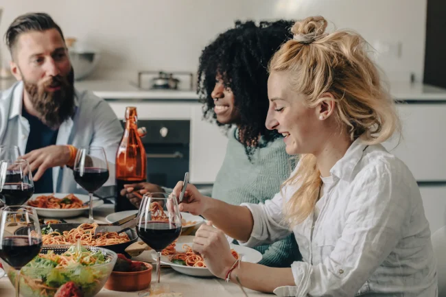 A group of friends laugh while eating a spaghetti-based meal together at home.