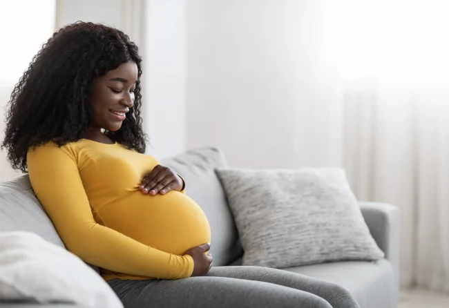 A pregnant woman in a yellow top sits on a sofa and cradles her bump happily.