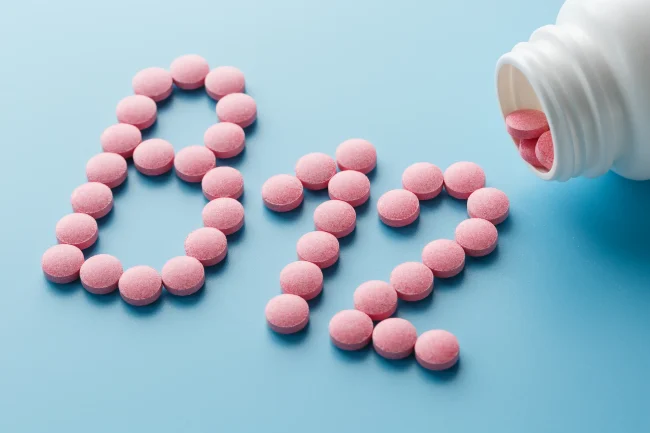 Small, round, pink tablets spill onto a blue surface, spelling out ‘B12’. Small, round, pink tablets spill onto a blue surface, spelling out ‘B12’.